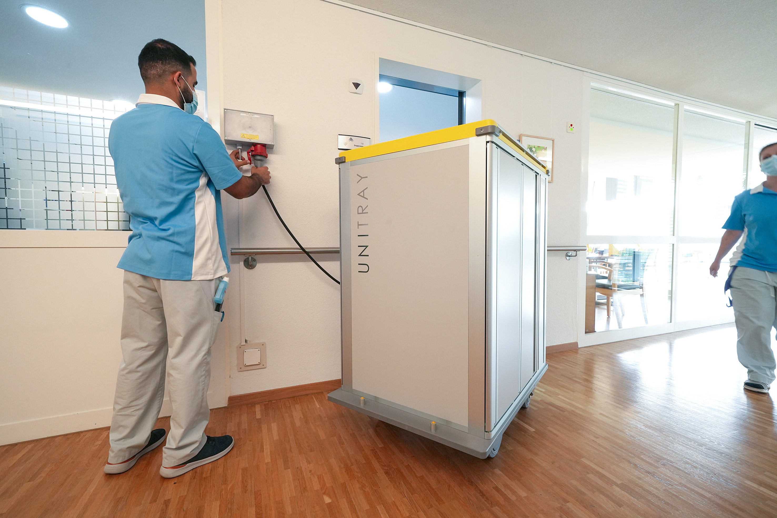 Auf den Etagen, wo wegen einer Grippewelle vorübergehend Maskenpflicht herrschte, werden die Wagen an den Strom angeschlossen und die Speisen mittels Heissluft rechtzeitig heiss und servierbereit gemacht.