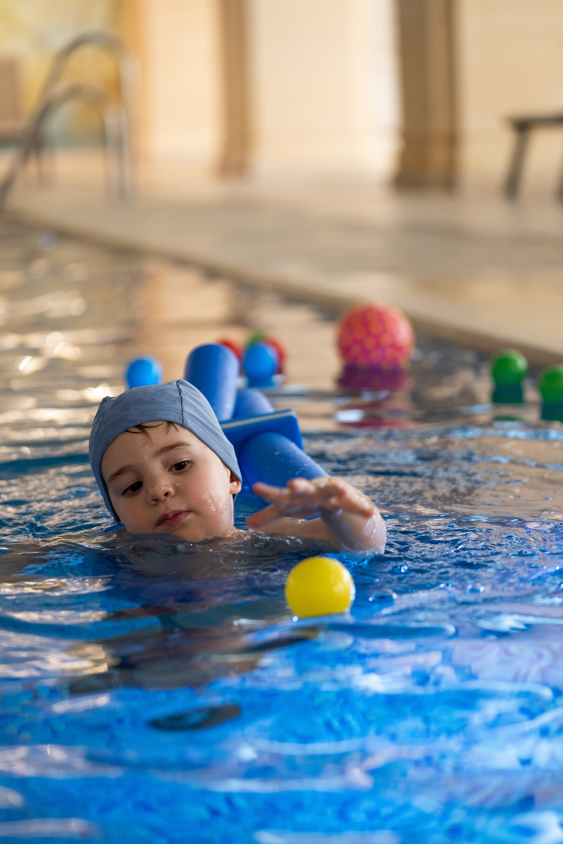 Child swimming with floatation device and playing with ball in indoor pool