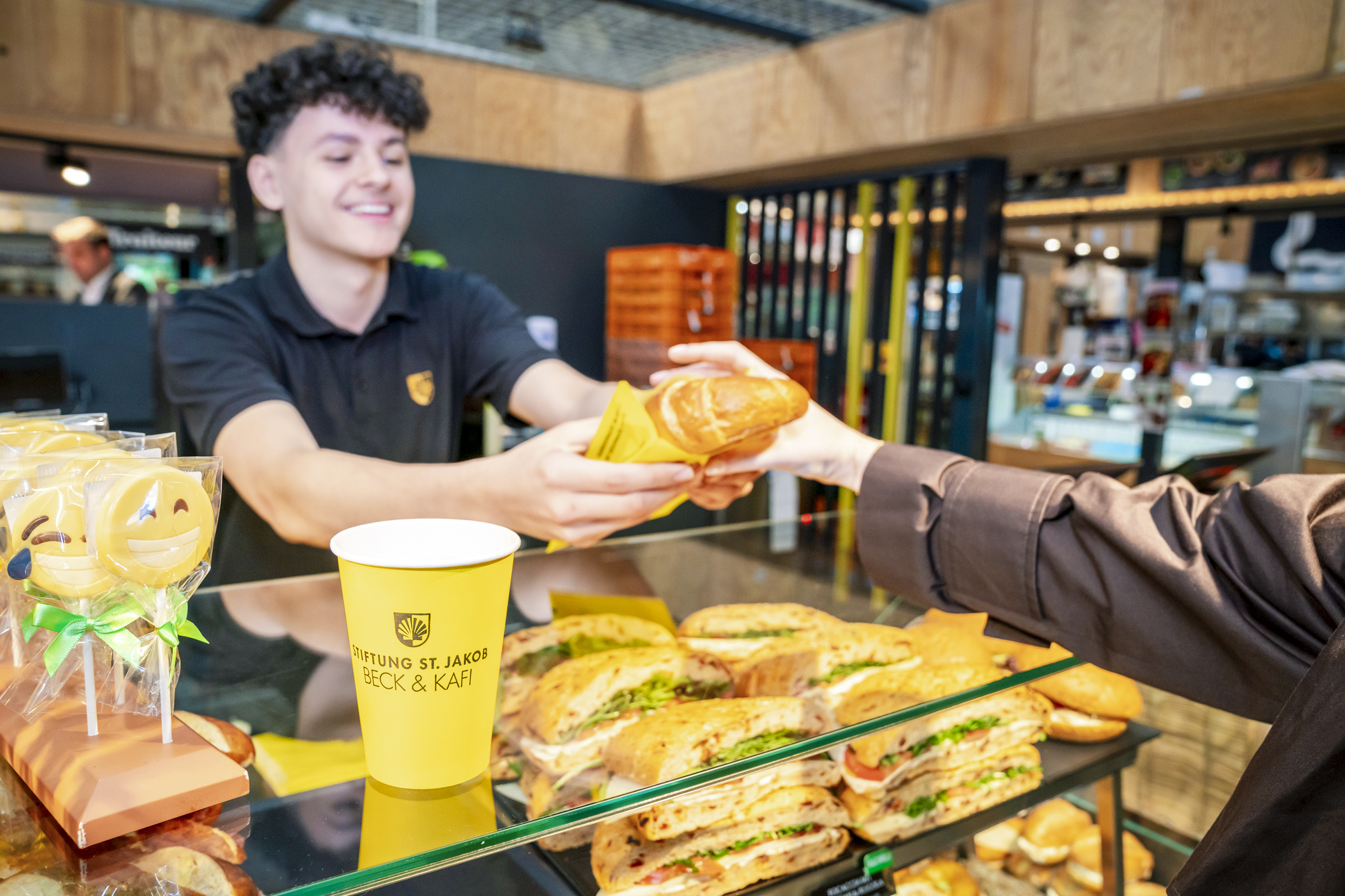 Freundlicher Service: ein Mitarbeiter reicht einer Kundin einen Kaffee to go und ein ofenfrisches Gipfeli.
