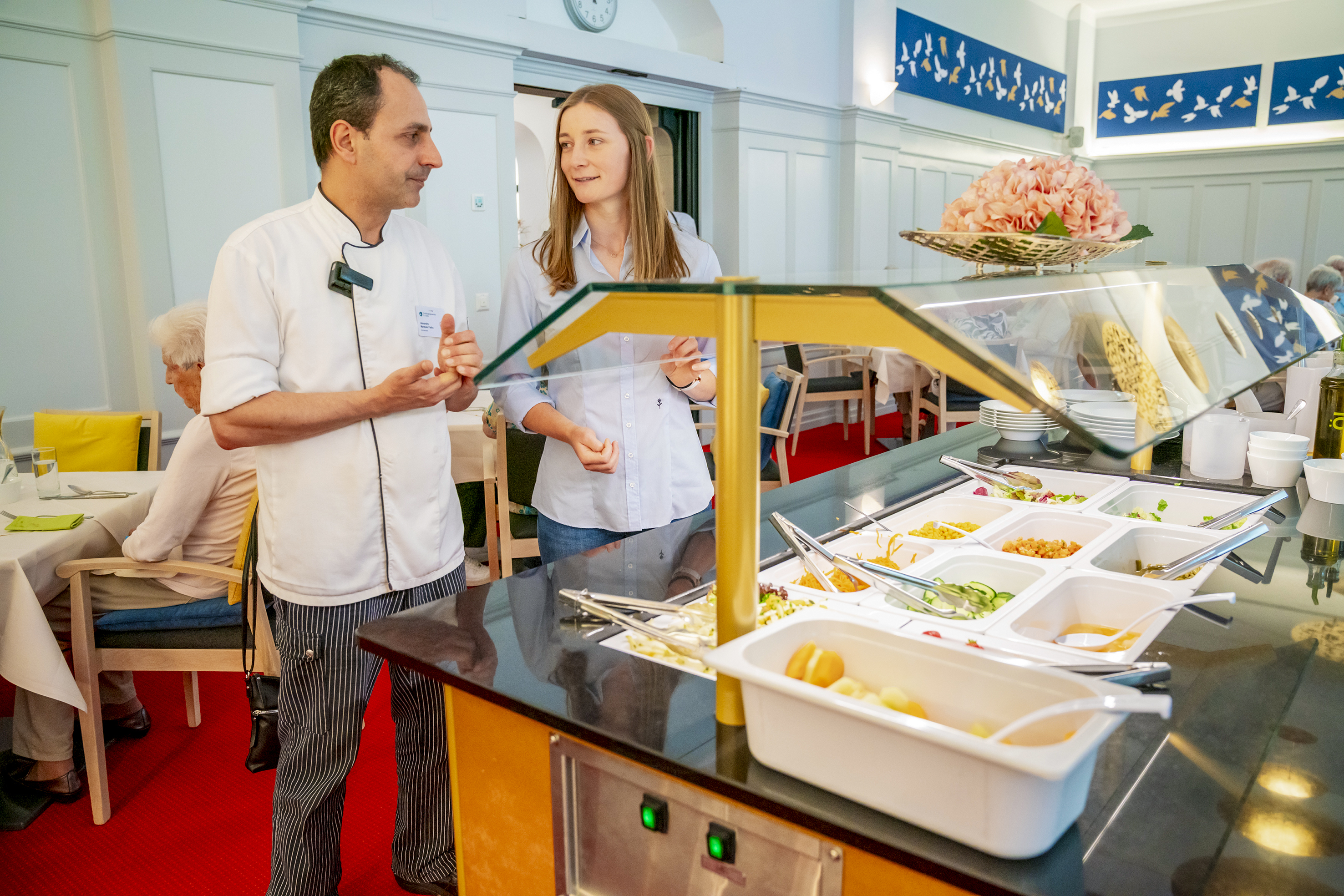 Küchenchef Alex Marques Fialho und Julia Menet von megaPlus tauschen sich aus: Dank unterfahrbarer Granitabdeckung können auch Bewohnende mit Rollator oder im Rollstuhl bequem ans Salatbuffet.